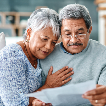 An elderly couple sitting down and reading documents, with a woman’s hand on her partner’s chest.